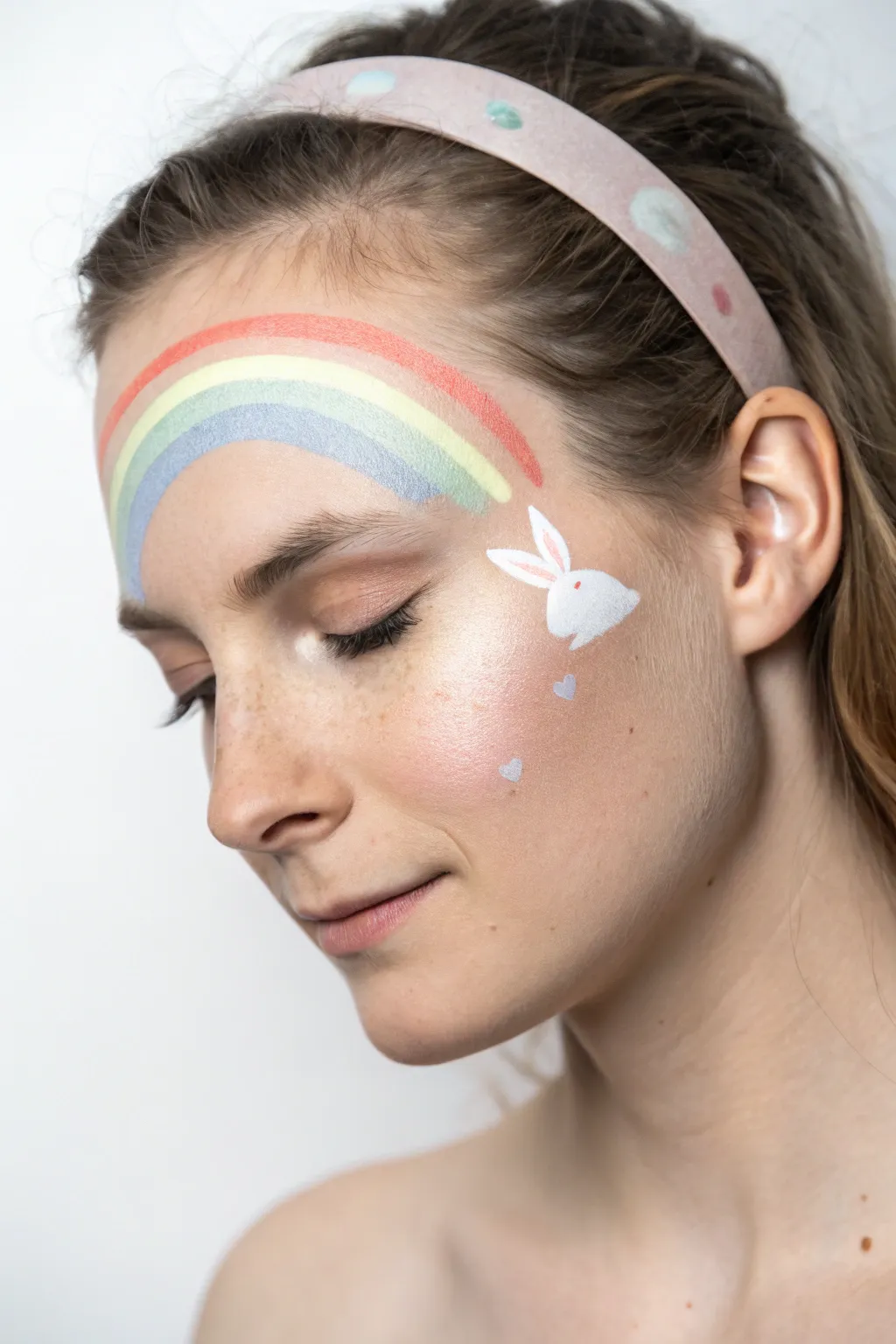 Soft pastel rainbow arch face paint with minimalist bunny silhouette and tiny Easter eggs