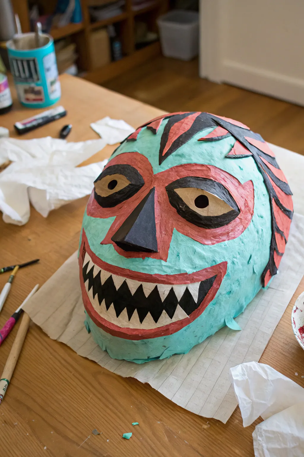 Paper mache monster mask with exaggerated eyes and grin, shown half raw strips, half boldly painted