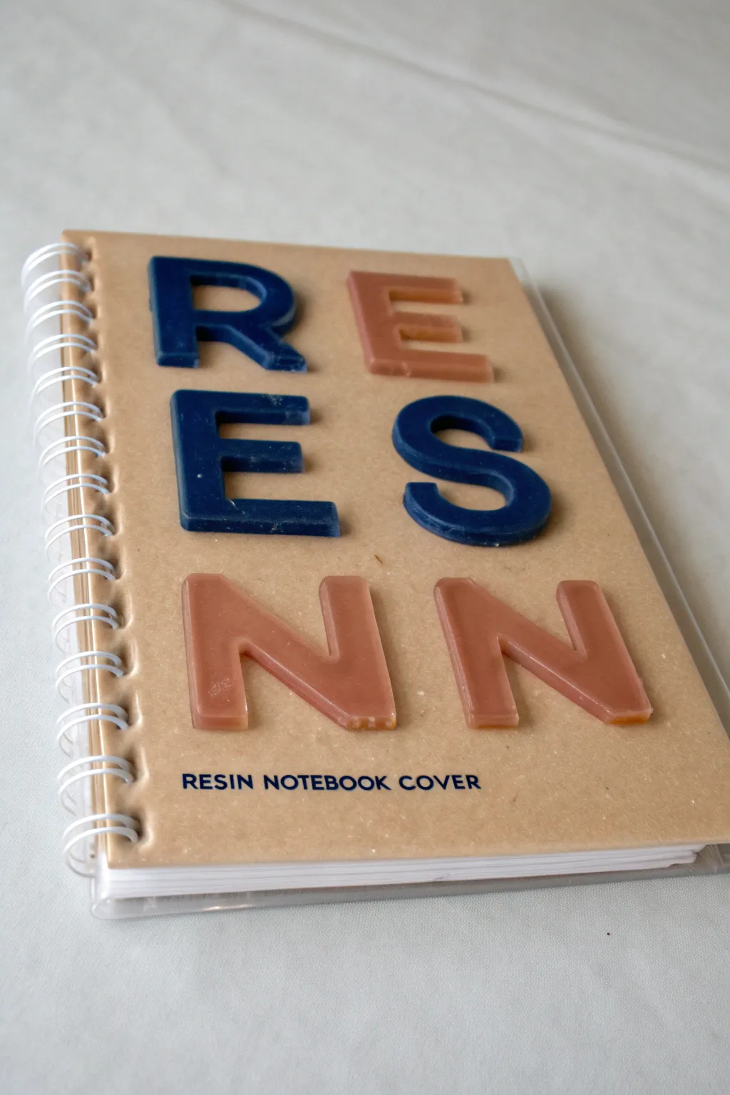 Minimal resin notebook cover with raised 3D letter embeds under glossy clear topcoat, bold contrast