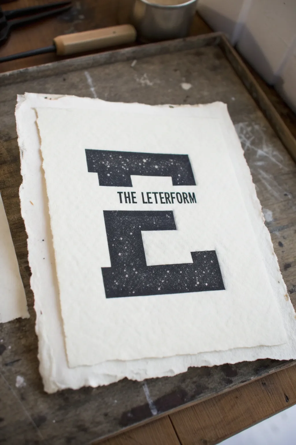 Minimal negative space lettering: crisp white letter cut from rich black ink with handmade speckles.