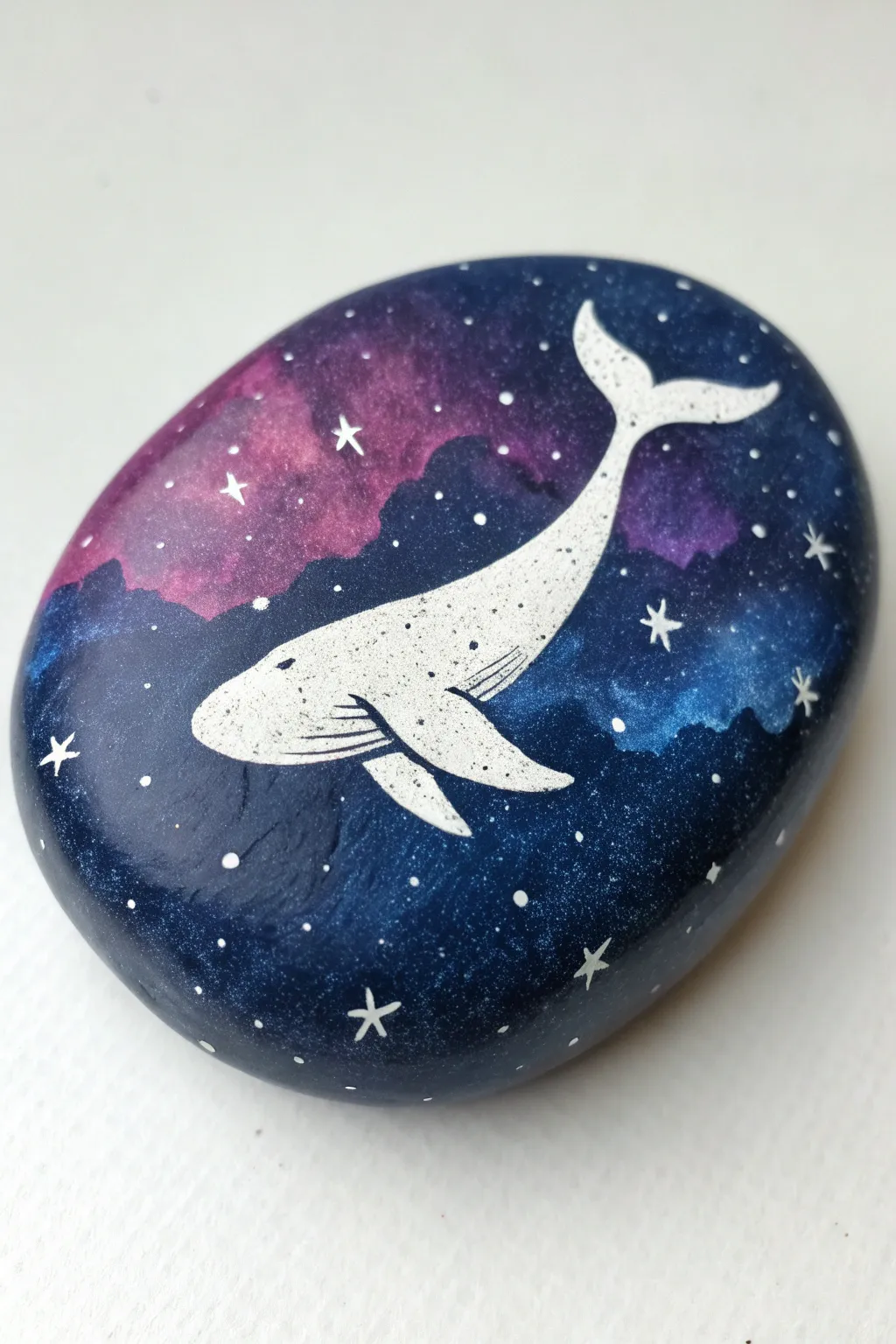 Paint a cosmic whale on a stone canvas with moody galaxy blues, purples, and bright stars