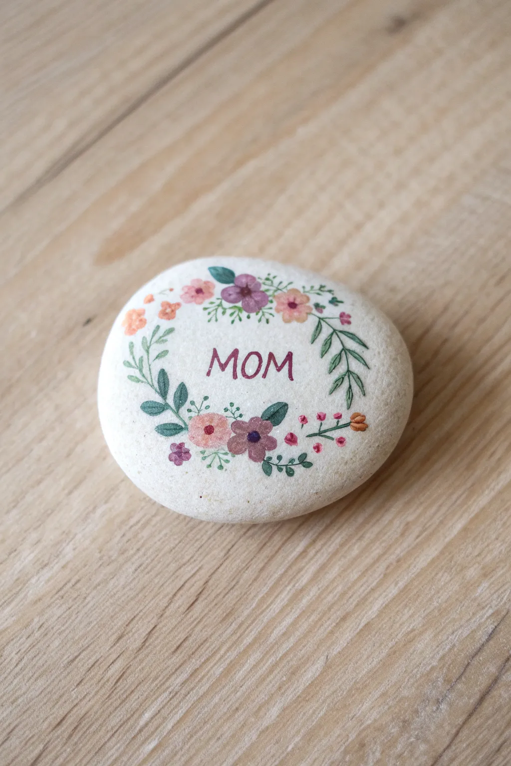 Simple Mothers Day rock art: floral wreath framing Mom on an oval stone, minimalist boho on wood.