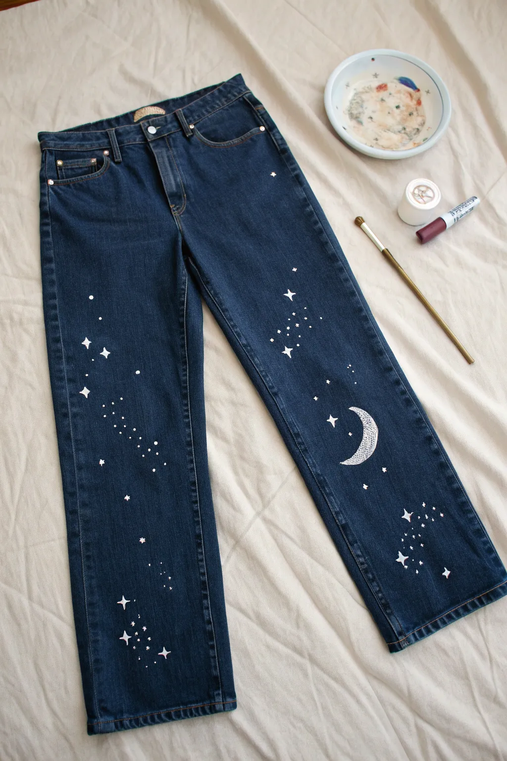 Celestial night-sky jeans laid flat with hand-painted stars and moons, simple and dreamy