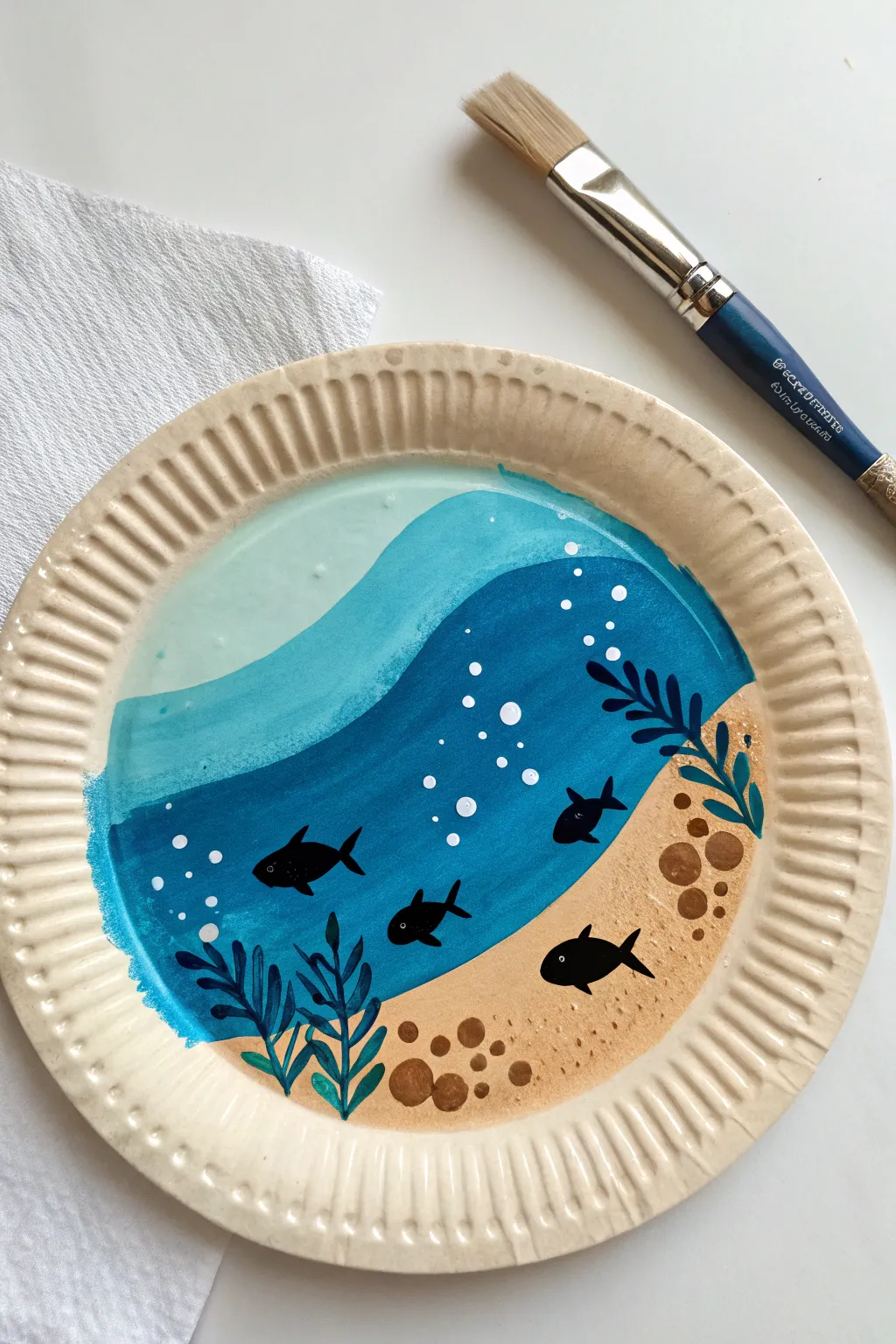 Underwater aquarium paper plate painting with layered blues, sand, and simple fish silhouettes