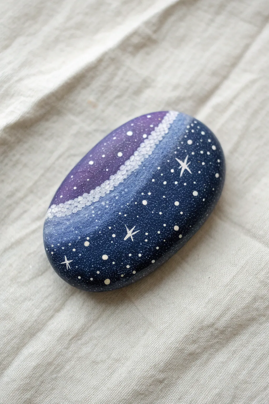 Paint a dreamy Milky Way sweep on a smooth oval stone, with stars clustered in the band.
