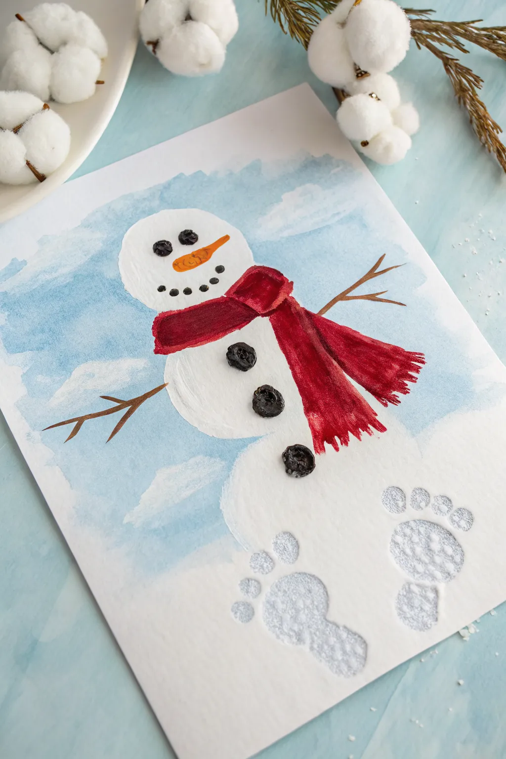 Snowman from baby footprints with a bold red scarf on an icy blue wash, simple and wintry