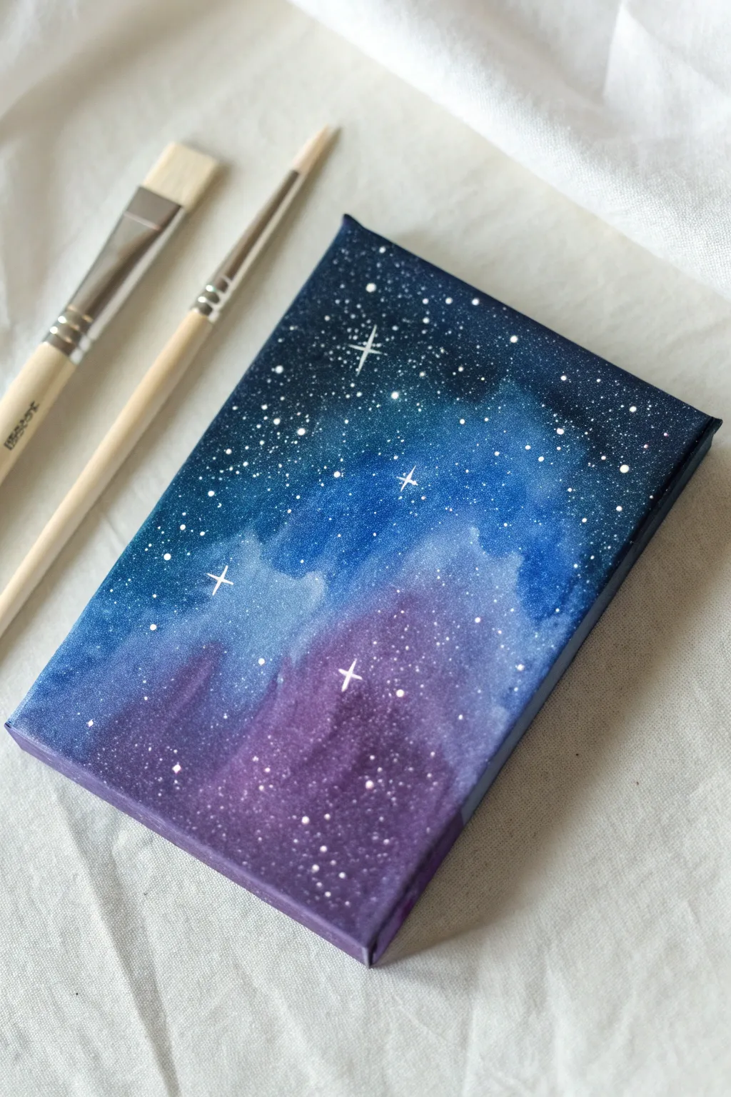 Paint a dreamy galaxy on a small canvas panel with splattered stars and a glowing nebula.