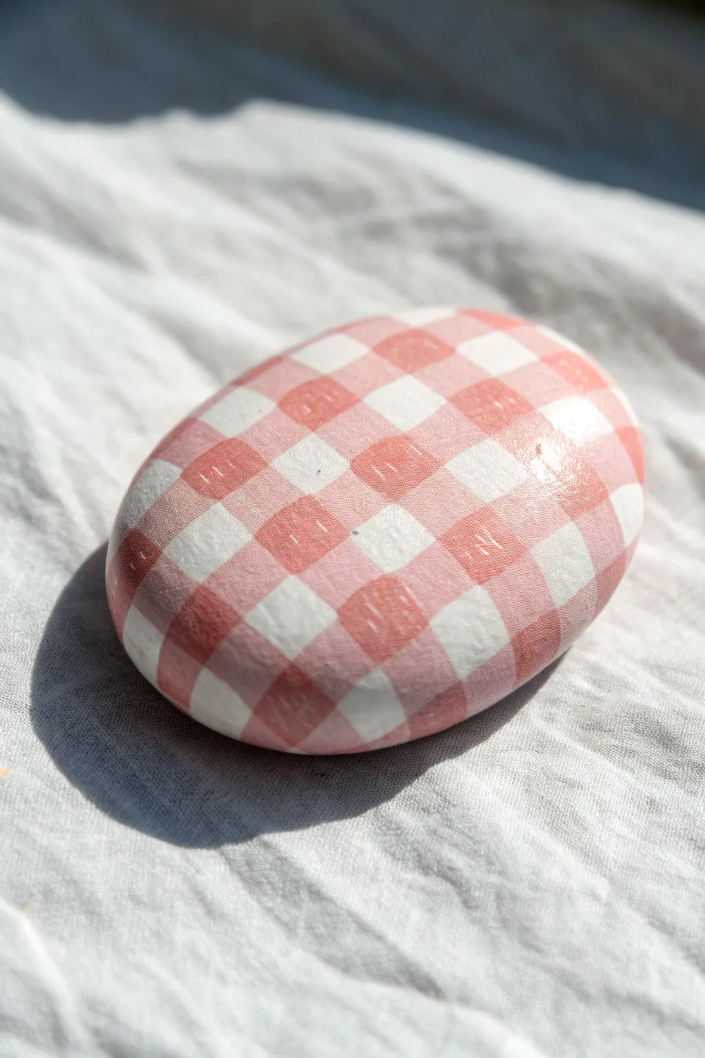 Blush pink-and-white gingham picnic rock, crisp preppy grid on a smooth oval stone.